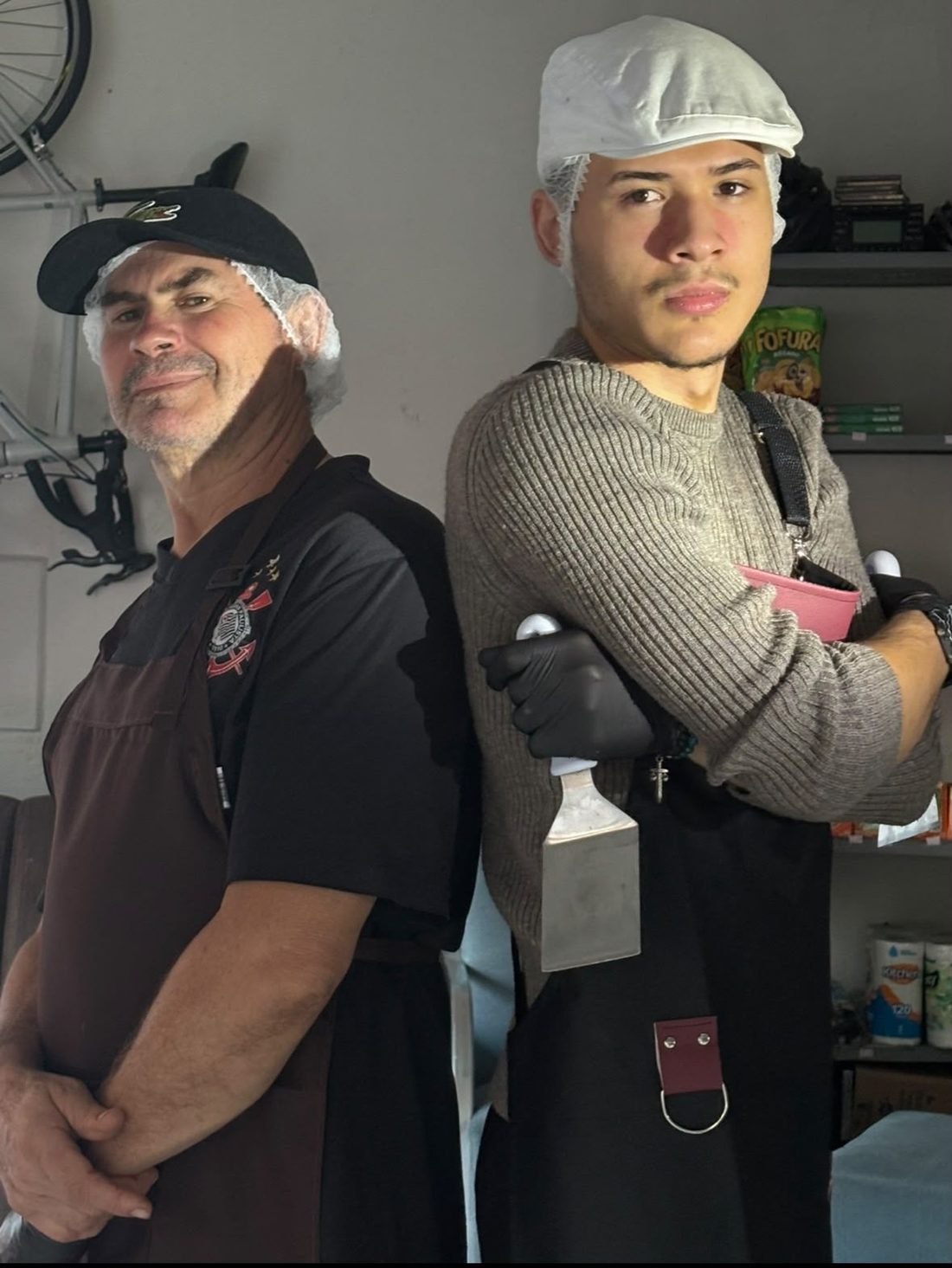 Pai e filho de touca e avental, costas com costas, na cozinha da Bruto Burguers