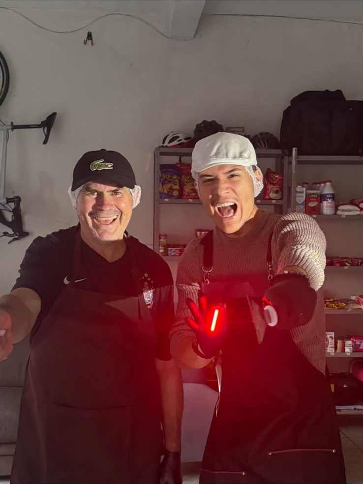 Pai e filho sorrindo e apontando para a câmera na cozinha, sob uma luz vermelha
