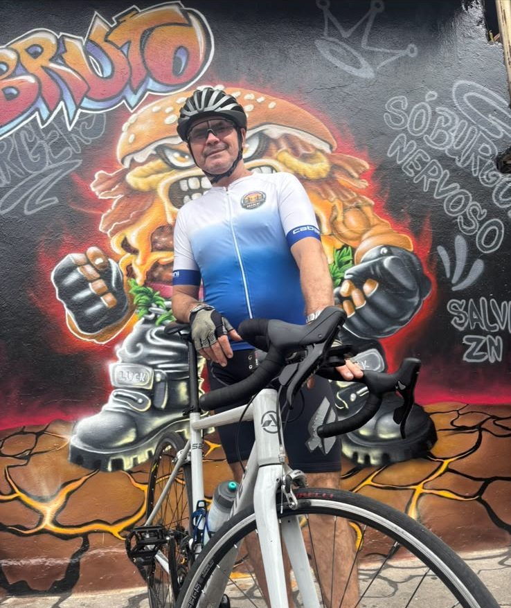 O pai, de roupa de ciclismo e capacete, com a bike diante do mural graffiti do Brutizila